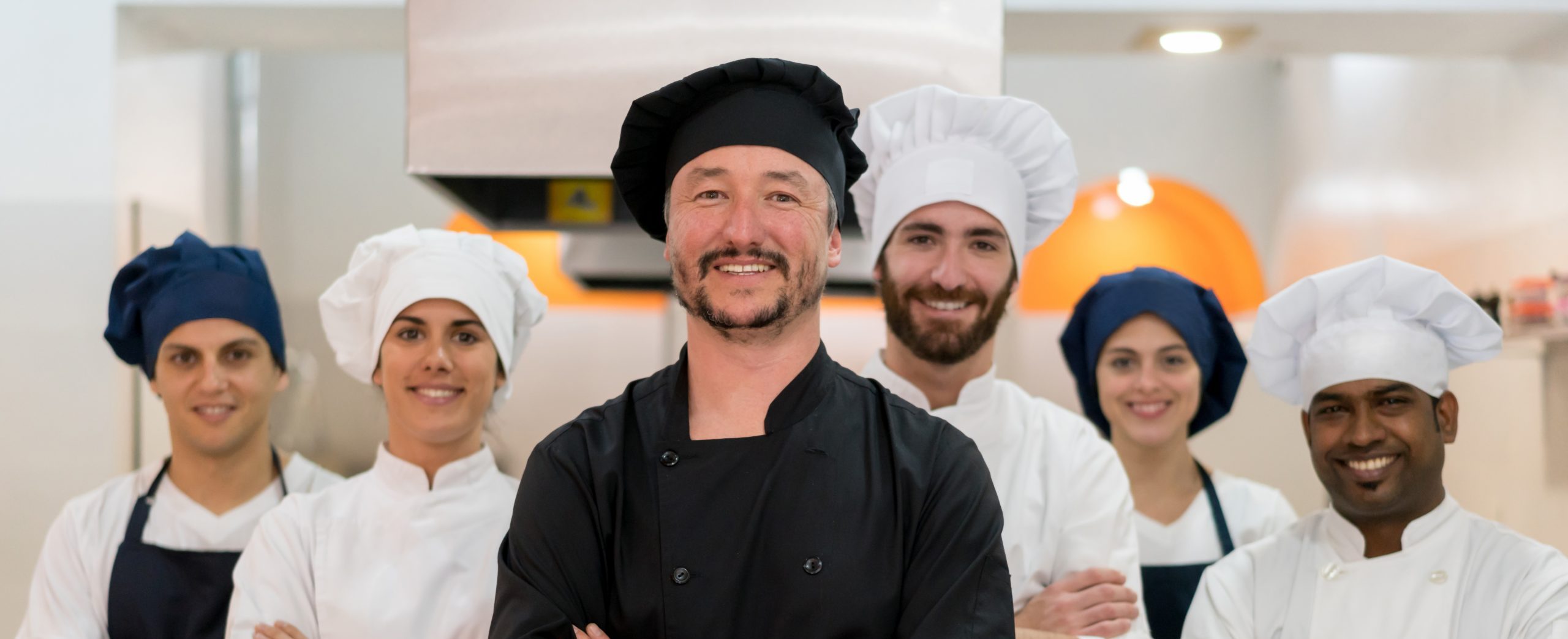 Proud multi ethnic team of chef, sous chefs and cooking assistants looking at camera smiling with arms crossed